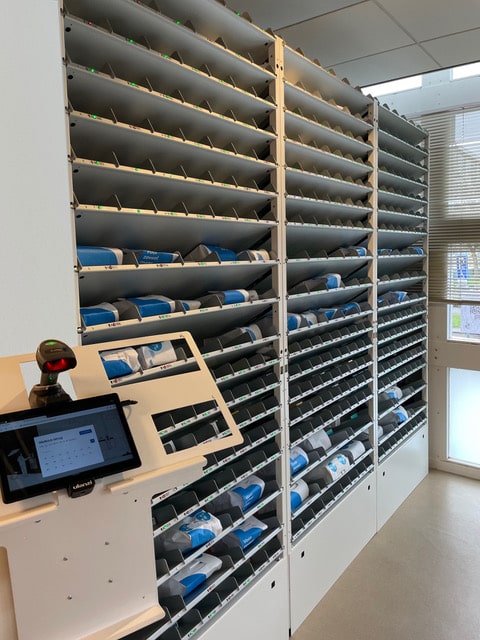 Binnenkant van een apotheek met een Pharmastore afhaalsysteem: meerdere smalle rekken gevuld met medicatiepakketjes en een touchscreen bedieningspaneel met scanner op de voorgrond.