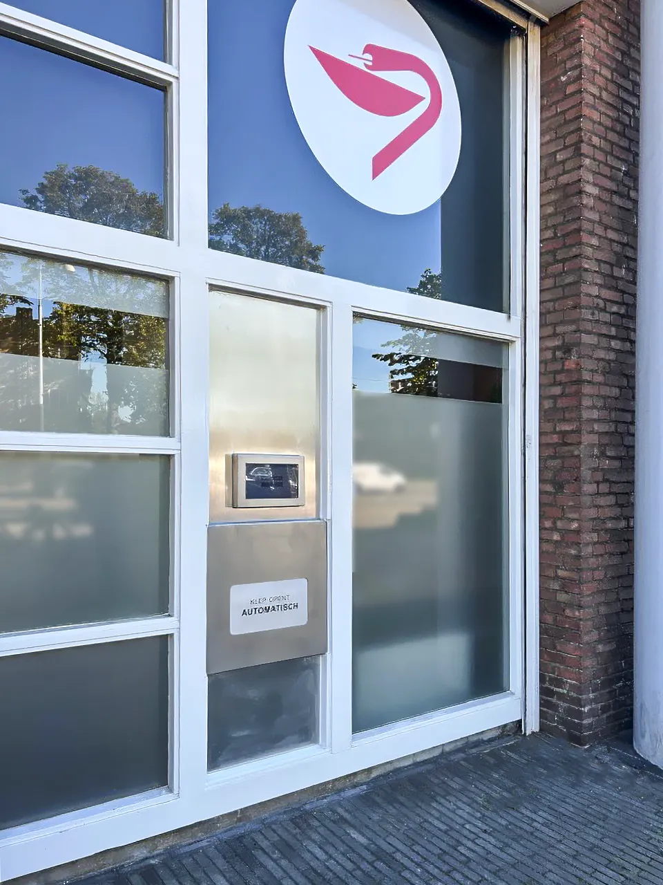 Buitenkant van een gebouw met grote ramen, een logo van een rode zwaan in een cirkel op het raam, en een metalen brievenbus of automaat met het opschrift 'Klep opent automatisch.