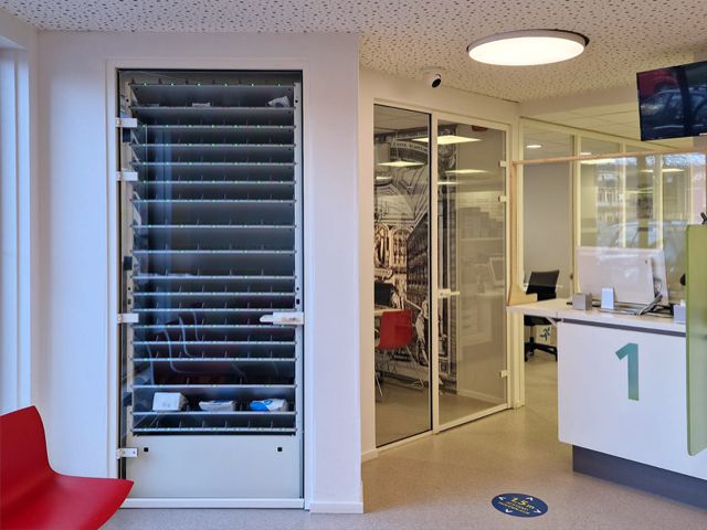 Binnenzijde van een apotheek met een geavanceerd Pharmastore afhaalsysteem, bestaande uit meerdere smalle rekken gevuld met medicatiepakketjes, en een touchscreen bedieningspaneel met scanner op de voorgrond