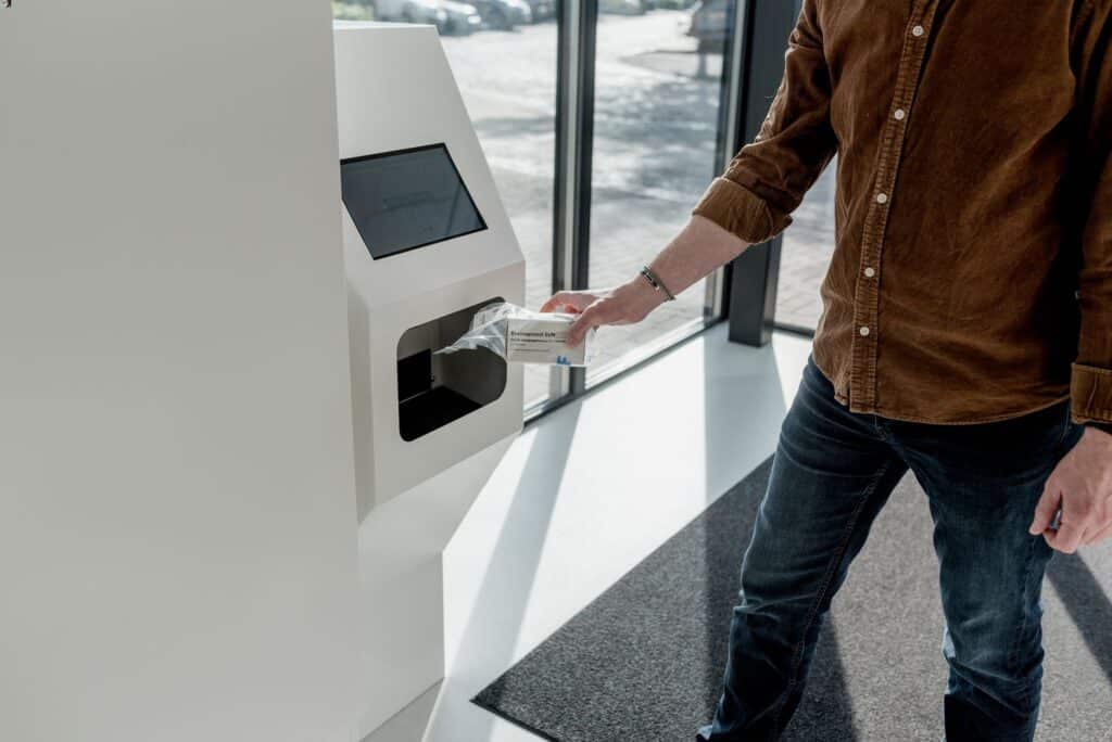 Een persoon haalt een medicijnpakketje uit een witte medicatie-uitgifteautomaat met touchscreen, geplaatst in een moderne ruimte met grote ramen en veel daglicht. De persoon draagt een bruine blouse en jeans en staat op een donkere mat bij de automaat.
