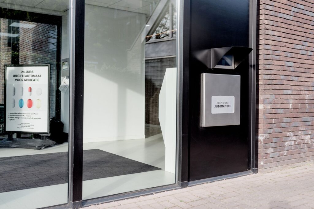Een moderne gevel van een apotheek met een 24-uurs medicatie-uitgifteautomaat. Binnen in de wachtruimte is een poster zichtbaar die uitleg geeft over de automatische medicatie-uitgifte.