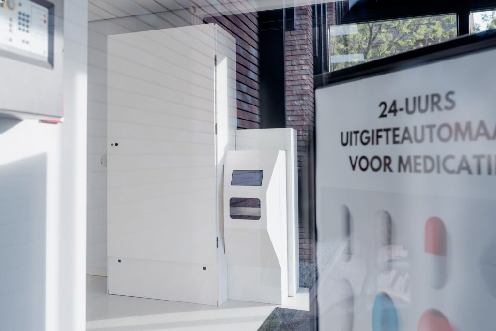 Binnenaanzicht van een apotheek met een witte, moderne medicatie-uitgifte-unit achter een glazen wand, zichtbaar naast een poster over de 24-uurs uitgifteservice.