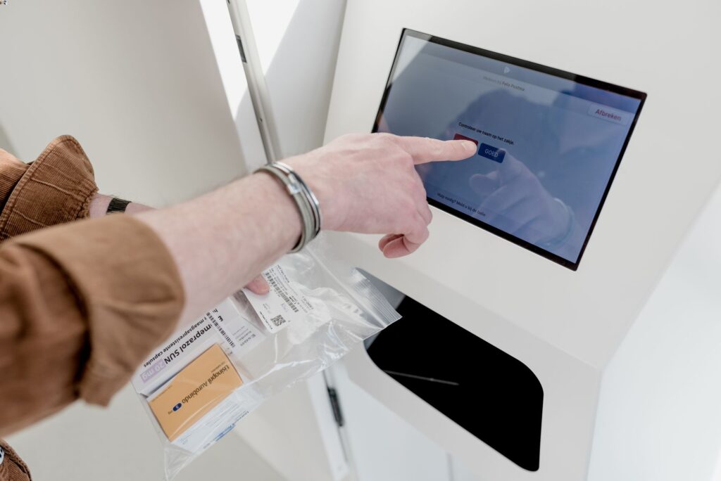 Een persoon bedient een touchscreen van een medicatie-uitgifteautomaat en bevestigt via de knop 'GOED' de naam op een ontvangen medicatiepakketje. De persoon draagt een bruine blouse en houdt een transparant zakje met medicijnen vast. De omgeving oogt licht en klinisch.