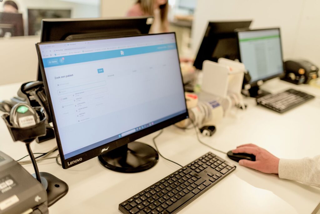 Een bureaublad met een Lenovo-computerscherm waarop een zoekpagina van een farmaceutisch pakketvolgsysteem zichtbaar is. Op de achtergrond staan meerdere werkplekken met schermen en apparatuur zoals scanners en labelprinters. Een hand bestuurt een computermuis, terwijl op de voorgrond een barcodelezer klaarstaat. De setting is een moderne en georganiseerde kantoor- of apotheekomgeving.