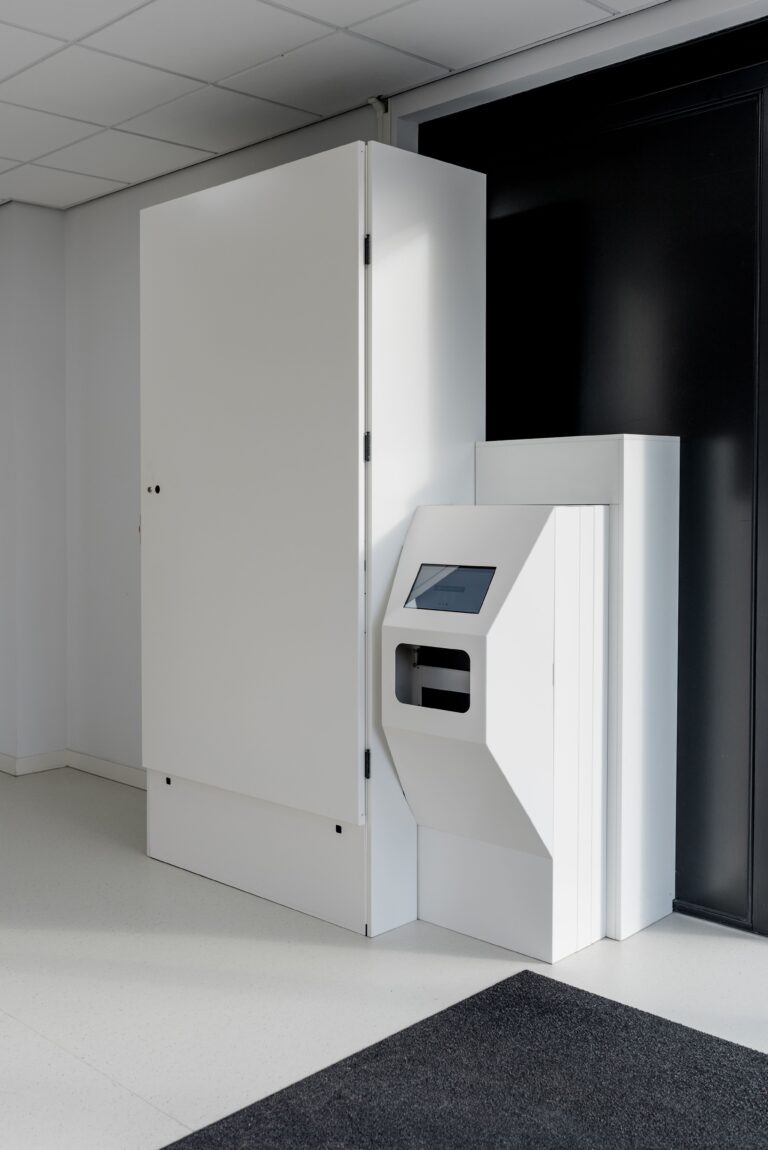 Een witte medicatie-uitgifteautomaat in een moderne, minimalistische ruimte met lichte vloeren en muren. De automaat bestaat uit een groot wit opbergdeel en een kleiner interactief paneel met een touchscreen en een opening voor medicatie-uitgifte. De ruimte oogt schoon en professioneel, met een donker vloerkleed voor de automaat.