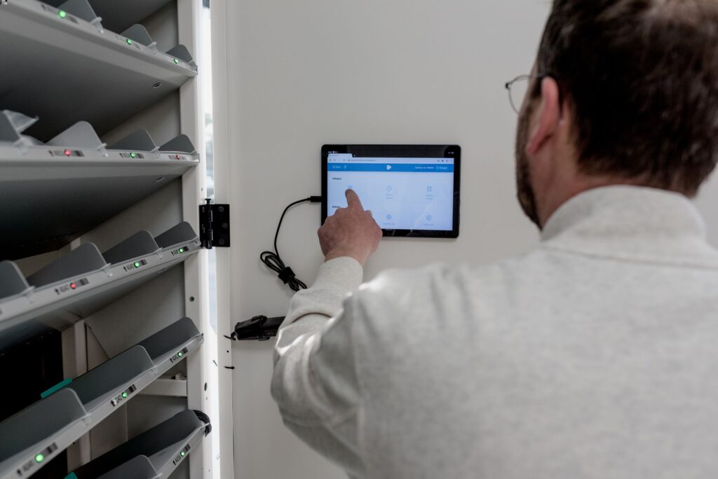 Een man bedient een aan de muur gemonteerd touchscreen binnen een geautomatiseerd medicatieopslagsysteem. Naast hem zijn meerdere medicijnvakken zichtbaar, gemarkeerd met groene en rode signaallampjes. De ruimte is licht en functioneel ingericht.
