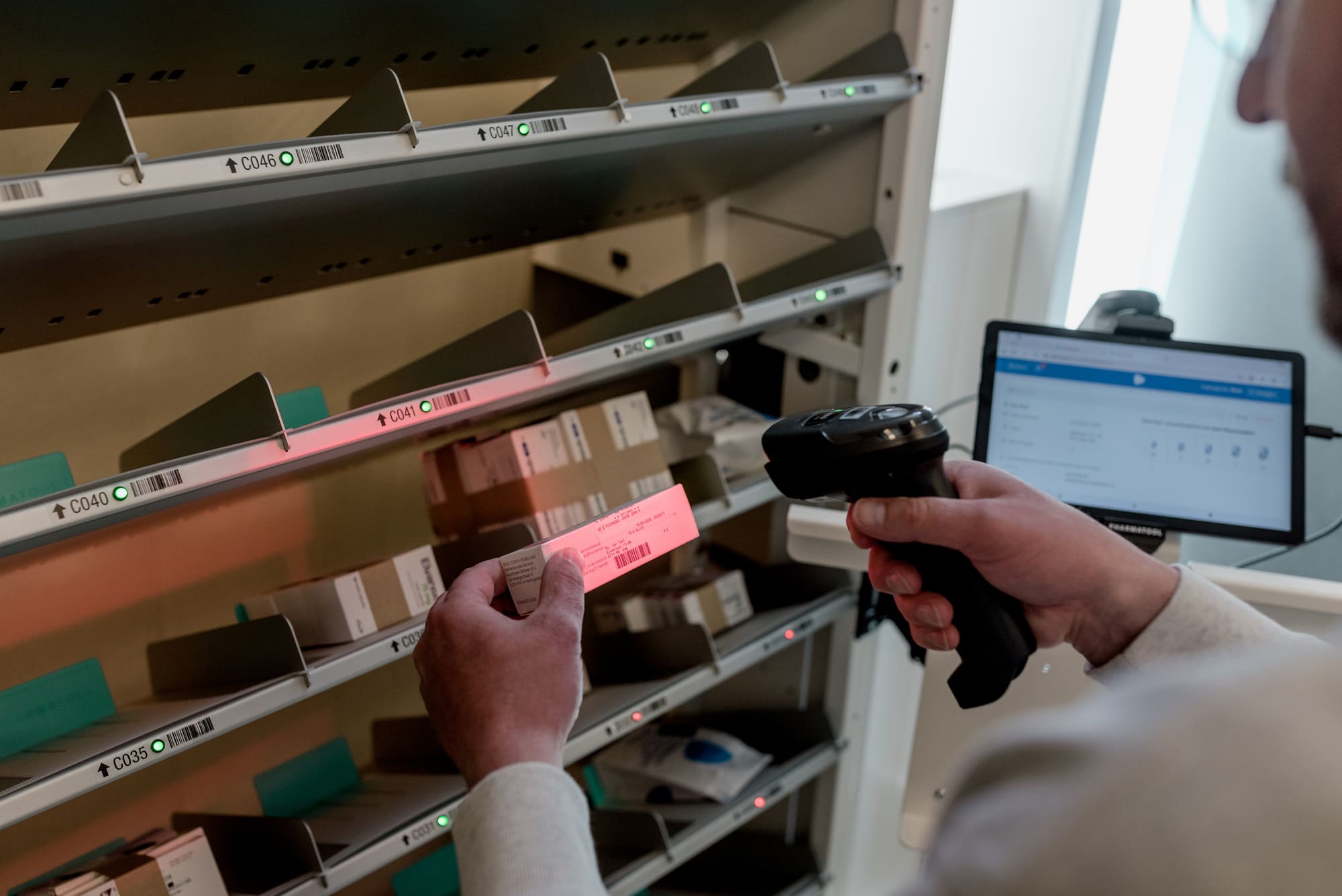 Close-up van een geautomatiseerd apotheekrek: een medewerker scant een rood verlicht barcodestrookje met een handheld scanner, terwijl groene LED-lampjes de juiste vaklocaties aangeven en een laptop met apotheeksoftware op de achtergrond meedraait.