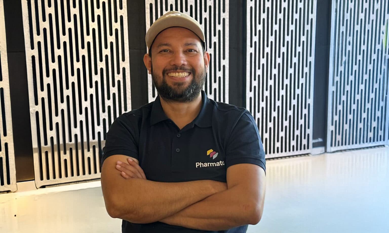 Lachende man met pet en donkerblauw Pharmatool-polo staat met gekruiste armen voor een moderne wand met verticale sleuven.