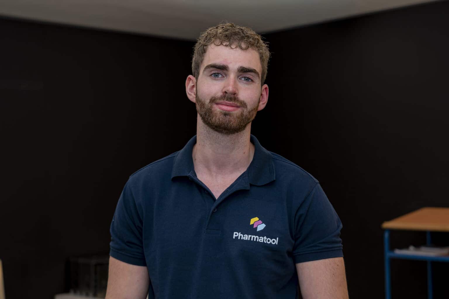 Een jonge man met krullend haar en een baard draagt een donkerblauw poloshirt met het logo van Pharmatool, terwijl hij recht in de camera kijkt tegen een donkere achtergrond.